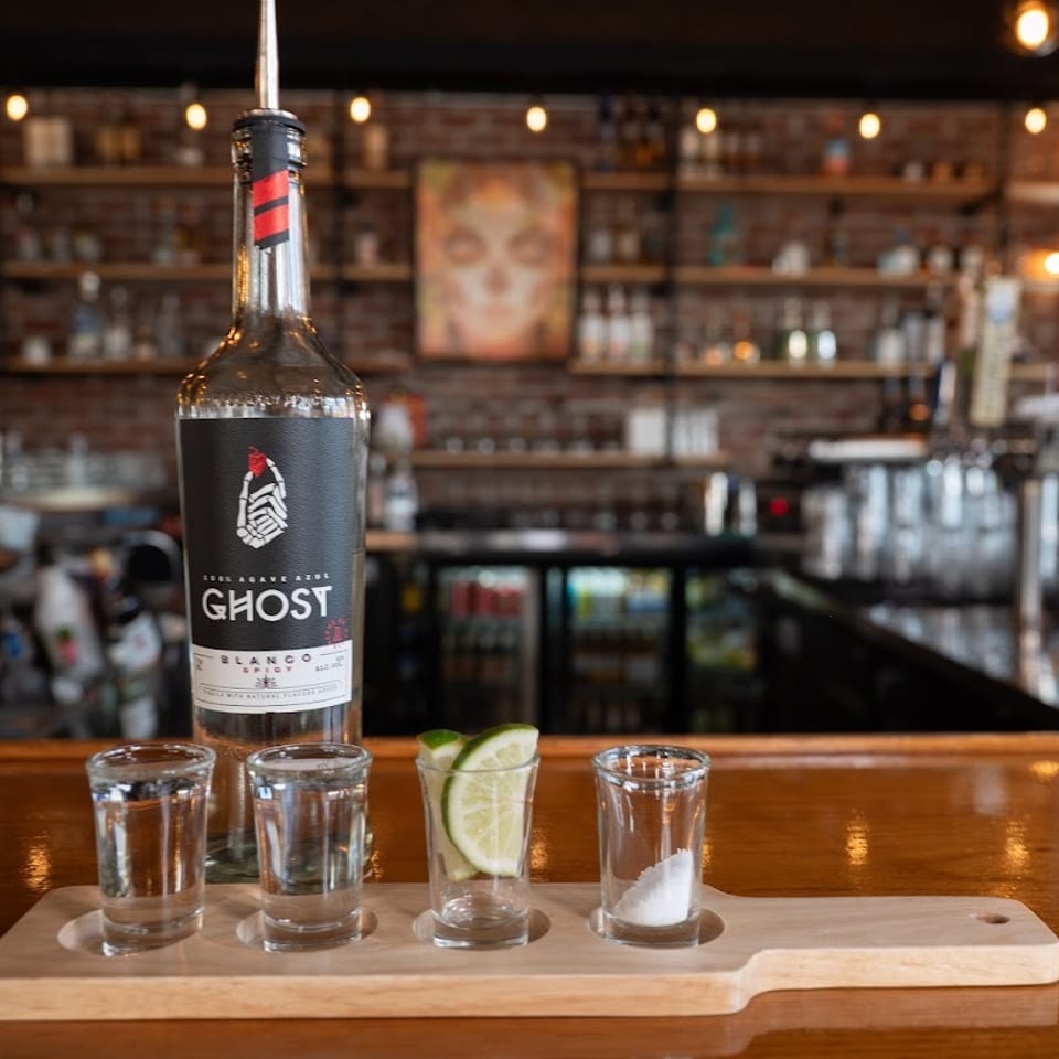 Ghost Blanco tequila bottle with tasting glasses, lime wedges, and salt on a wooden bar in a warmly lit restaurant.