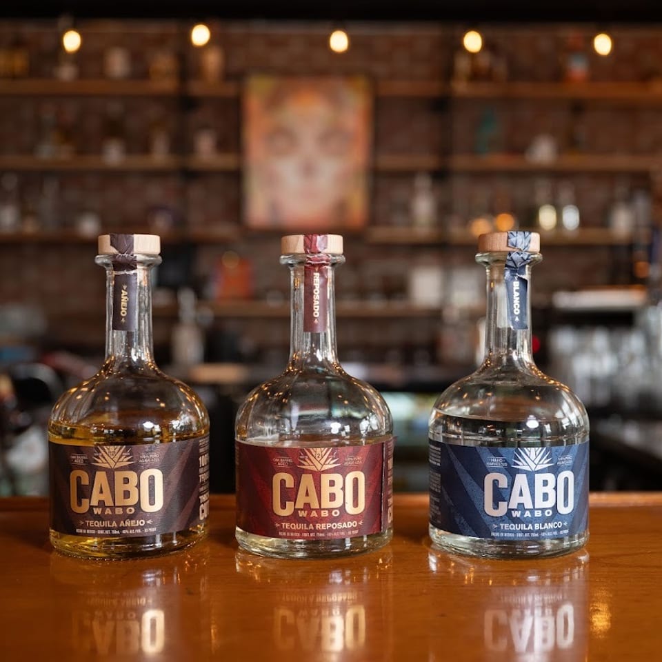Three Cabo Wabo tequila bottles—Añejo, Reposado, and Blanco—displayed on a polished bar beneath warm string lights.