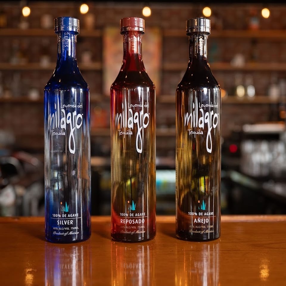 Three Milagro tequila bottles—Silver, Reposado, and Añejo—displayed on a polished bar in a warmly lit restaurant.
