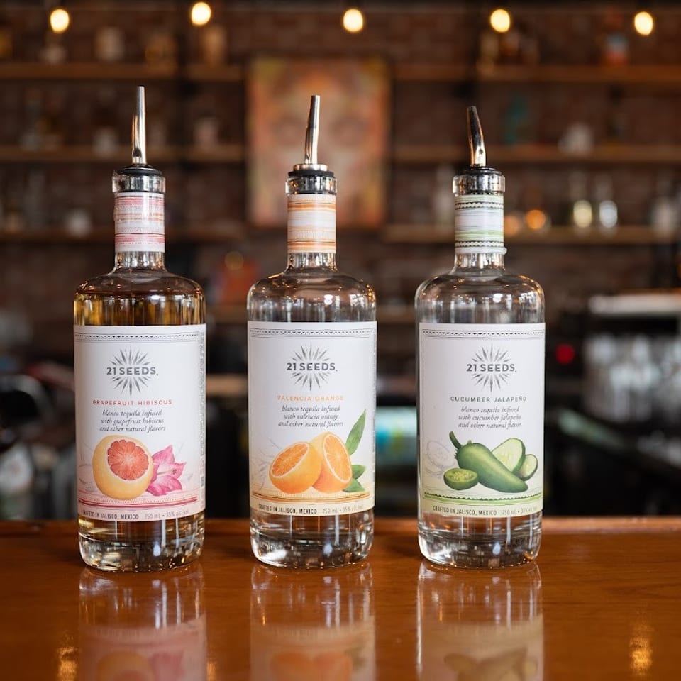 Three 21 Seeds flavored tequila bottles—grapefruit hibiscus, Valencia orange, and cucumber jalapeño—stand on a warmly lit bar.