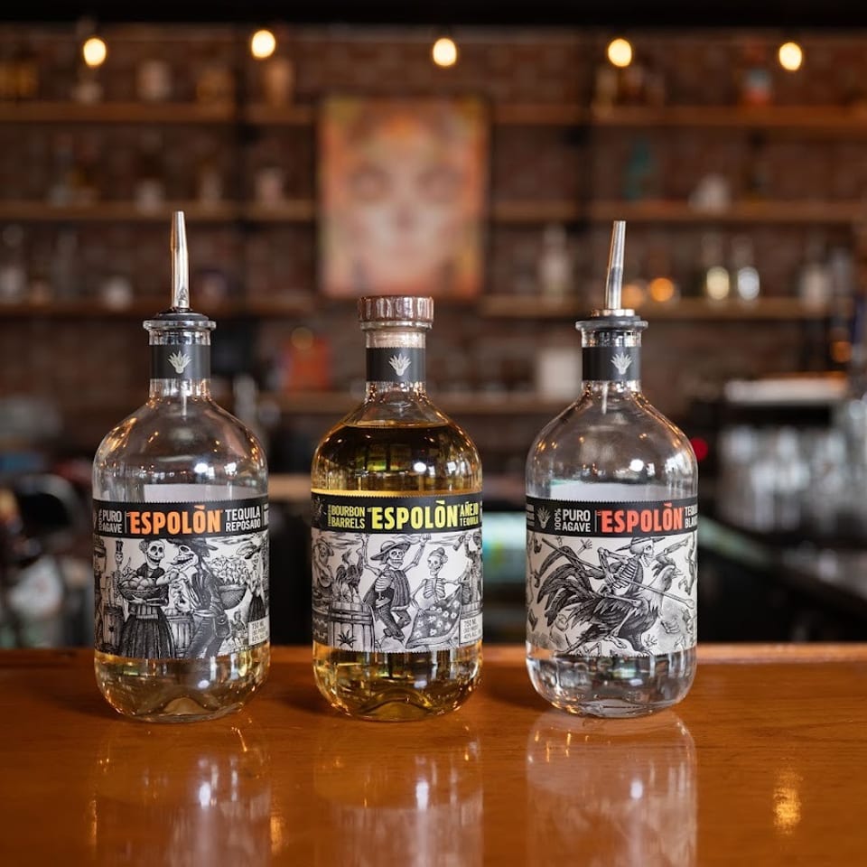 Three Espolón tequila bottles displayed on a polished wooden bar, with a warmly lit brick-walled restaurant behind them.
